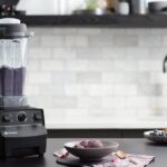 Licuadora vitamix 5200 sobre una mesa negra de una cocina moderna. Dentro del vaso de la licuadora se observa una bebida de color violeta. A sus cotados se ven ingredientes frescos como aguacate u arándanos.