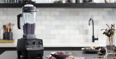 Licuadora vitamix 5200 sobre una mesa negra de una cocina moderna. Dentro del vaso de la licuadora se observa una bebida de color violeta. A sus cotados se ven ingredientes frescos como aguacate u arándanos.