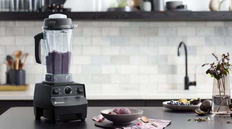 Licuadora vitamix 5200 sobre una mesa negra de una cocina moderna. Dentro del vaso de la licuadora se observa una bebida de color violeta. A sus cotados se ven ingredientes frescos como aguacate u arándanos.
