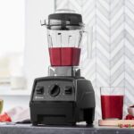 Licuadora Vitamix E320 sobre una mesa en una cocina moderna. Dentro del vaso de la licuadora se observa una malteada de color rojo. A sus costados se ven ingredientes frescos como, sandía, frambuesas y limón.