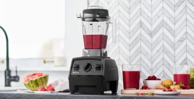 Licuadora Vitamix E320 sobre una mesa en una cocina moderna. Dentro del vaso de la licuadora se observa una malteada de color rojo. A sus costados se ven ingredientes frescos como, sandía, frambuesas y limón.