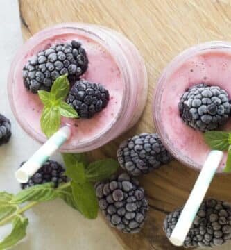 Dos licuados de frutos rojos con leche en un vaso de vidrio con popote cada uno. Están sobre una tabla de madera en una mesa blanca y alrededor se encuentran moras frescas.