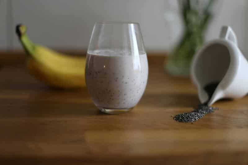Licuado de plátano con semillas de chía en un vaso de vidrio. A sus costados se ve un plátano maduro y un tazón de semillas de chía derramadas. Todo sobre una mesa de madera.