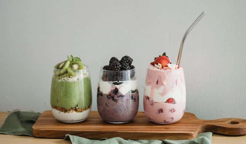 Tres vasos con licuado de frutas, con ingredientes frescos como kiwi, fresas y zarzamoras. Todo sobre una tabla de madera en una mesa.