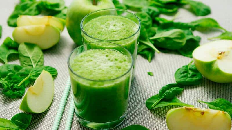 Dos licuados verdes en el centro, en vasos de vidrio pequeños. A sus costados hay ingredientes como espinacas y manzanas cortadas en gajos. Todo sobre un mantel de color blanco.
